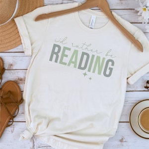 May include: A white t-shirt with the text "i'd rather be READING" in green and grey lettering. The shirt is on a hanger and has a knot tied at the bottom.