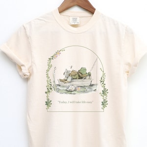 May include: A cream colored t-shirt with a green frog illustration. The frogs are sitting in a boat on a lily pad. The text "Today, I will take life easy" is printed below the illustration.