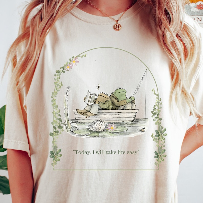 May include: A white t-shirt with a green illustration of two frogs in a boat. The frogs are reading a book and fishing. The text "Today, I will take life easy" is printed below the illustration.