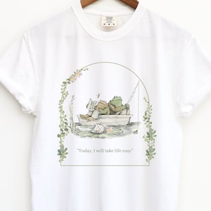 May include: White t-shirt with a green frog illustration. The frogs are in a boat with a fishing rod and a book. The text "Today, I will take life easy" is printed below the illustration.