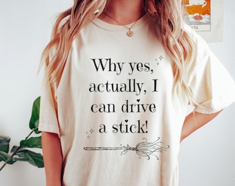 May include: Cream-colored t-shirt with the text "Why yes, actually, I can drive a stick!" in black. A black broom graphic is below the text, with two small star graphics on either side.