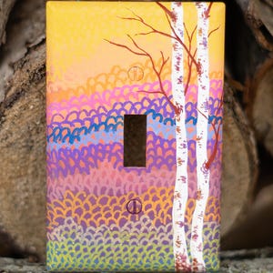 Könnte beinhalten: Handbemalte Lichtschalterabdeckung mit einem farbenfrohen Landschaftsdesign. Das Kunstwerk zeigt einen gelben Himmel, einen Farbverlauf aus Orange, Rosa, Blau und Lila sowie weiße Birken mit roten Ästen. Die Abdeckung hat eine einzelne Schalteröffnung.