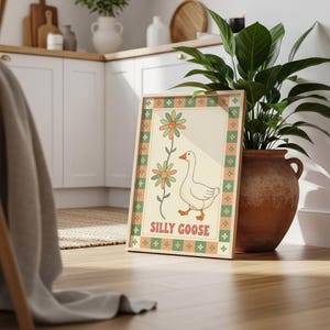 Może przedstawiać: Oprawiony wydruk z ilustracją gęsi i napisem "Silly Goose". Retro design wykorzystuje pomarańczowe, zielone i beżowe odcienie. Wydruk stoi na drewnianej podłodze, obok rośliny w terakotowej donicy.