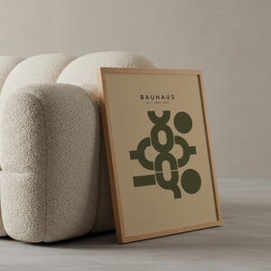 Puede incluir: Un póster enmarcado de Bauhaus con fondo beige, diseño abstracto verde oscuro y texto "BAUHAUS" y "WEIL SEPT 1923". El póster está apoyado contra un sofá crema redondeado.