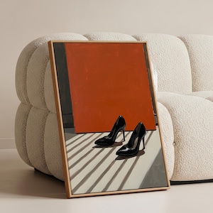 Può includere: Un'opera d'arte incorniciata con un design astratto arancione e grigio, con un paio di scarpe con tacco nere lucide davanti. L'opera d'arte è appoggiata a un divano arrotondato color crema.