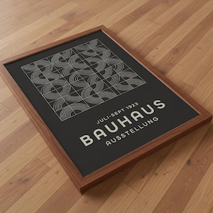 Puede incluir: Un póster enmarcado de la exposición Bauhaus. El póster presenta un diseño geométrico en blanco sobre un fondo negro. El texto "JULI-SEPT 1923 BAUHAUS AUSSTELLUNG" está impreso en blanco. El marco es de madera.