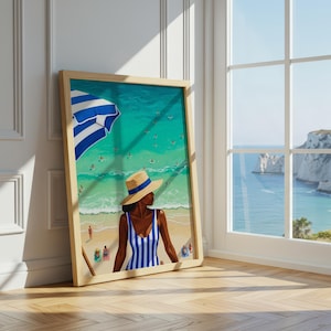 Può includere: Una stampa artistica incorniciata raffigura una donna in costume da bagno a righe blu e bianche e cappello di paglia su una spiaggia. È visibile un ombrellone a righe blu e bianche. L'opera d'arte è esposta su un pavimento di legno.
