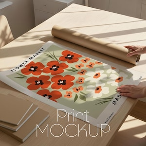 以下が含まれることがあります： オレンジと白の花が薄緑色の背景に描かれた花のプリントポスター。 「FLOWER MARKET」と「Print Collection」の文字が見えます。 茶色の丸められた紙管といくつかの茶色のノートもテーブルにあります。