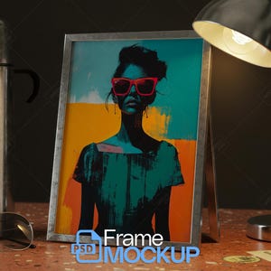 May include: A framed art print with a silver frame, depicting a woman in red sunglasses. The artwork uses teal, orange, and black. A silver lamp and a stainless steel thermos are in the background.