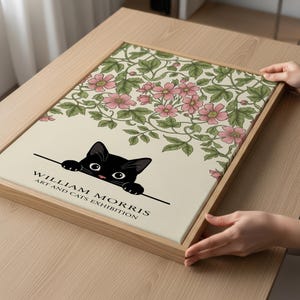 Op de afbeelding: Een ingelijste kunstprint met een zwarte kat en de tekst "WILLIAM MORRIS ART AND CATS EXHIBITION". De print toont roze bloemen en groene bladeren op een crèmekleurige achtergrond, in een licht houten frame, voor interieurdecoratie.