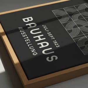 May include: A framed Bauhaus exhibition poster. The poster features the word "BAUHAUS" in large, silver letters, with "AUSSTELLUNG" below. The poster also includes the dates "Juli-Sept 1923" and a geometric design in the upper right corner. The frame is made of light-colored wood.