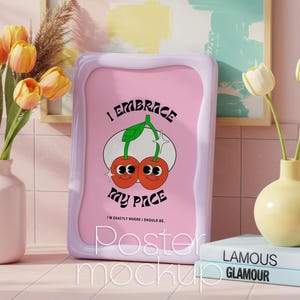 Puede incluir: Un póster rosa con un marco ondulado lila muestra el texto "I EMBRACE my PACE" y un diseño de cerezas de dibujos animados. El póster está apoyado sobre una superficie rosa claro, con un jarrón de tulipanes amarillos y una pila de libros cerca.
