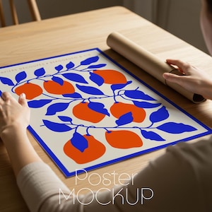 May include: A poster with a blue border featuring a design of orange fruit and blue leaves on a cream background. The poster is being held on a wooden table. A rolled-up poster is next to the poster.