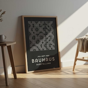 Puede incluir: Un póster enmarcado en blanco y negro de la exposición Bauhaus. El póster presenta un diseño geométrico de semicírculos y el texto "JULI-SEPT 1923 BAUHAUS AUSSTELLUNG". El póster está apoyado contra una pared blanca, junto a una mesa auxiliar de madera con un jarrón y una taza.