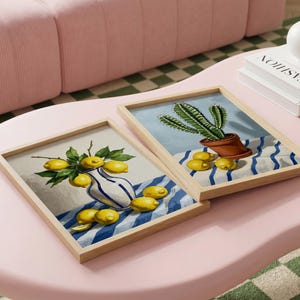 May include: Two framed art prints on a pink table. One features lemons in a vase, the other a cactus in a pot. Both are on blue and white striped fabric. The frames are light wood.