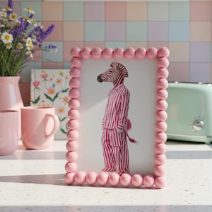 Peut inclure: Un cadre photo rose à perles avec une image fantaisiste d'un zèbre en tenue rayée. Le cadre est posé sur un comptoir avec des tasses roses, un vase de fleurs et un grille-pain vert menthe.