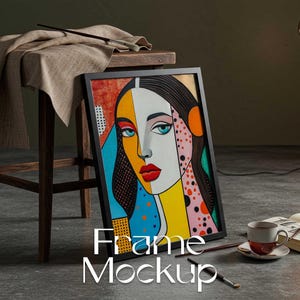 May include: A framed artwork with a colourful pop art style portrait of a woman with bold lines and vibrant colours. The artwork is propped against a wooden stool, with a coffee cup, book, and paintbrushes nearby. The text "Frame Mockup" is displayed at the bottom.
