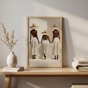 May include: A framed art print depicting three figures in white trousers and straw hats, set in a minimalist environment. The artwork is on a wooden table, with a vase of dried flowers and a folded blanket.