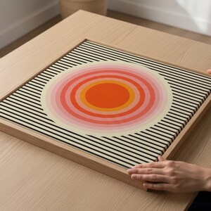 Peut inclure: Une impression d'art encadrée avec un design rétro. L'œuvre présente un motif circulaire central dans des tons orange, rose et crème, sur un fond de rayures noires et blanches. Le cadre est en bois de couleur claire.