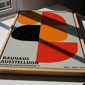 Könnte beinhalten: Gerahmtes Bauhaus-Ausstellungsplakat mit einem geometrischen Design in Orange, Rot, Schwarz und Grau. Das Plakat enthält den Text "BAUHAUS AUSSTELLUGN" und "JULI - SEPT 1923" in schwarzer Schrift. Der Rahmen ist aus hellem Holz.
