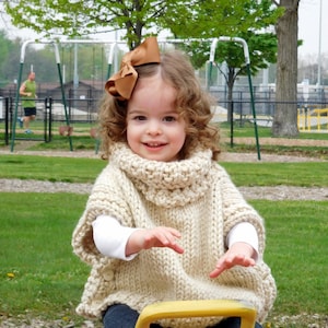 May include: A young girl wearing a cream-colored knitted sweater with a brown bow in her hair sits on a pink and yellow seesaw.