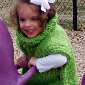 May include: A young girl wearing a green knitted sweater with a white bow in her hair is sitting on a purple playground equipment.
