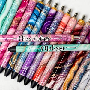 May include: A collection of personalized pens with glitter and marble-like designs. The pens are decorated with the names "Mrs. Adair" and "Melissa".