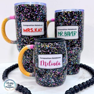 May include: Three composition notebook-themed tumblers and mugs. The black background is speckled with colorful paint flecks. Each mug has a label with a name: MRS. KAY, MR. BAKER, and Melinda. The mug handles are designed to look like pencils.