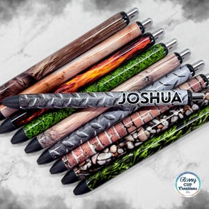 May include: A collection of ballpoint pens with various patterned designs, including wood grain, flames, grass, and brick. One pen features the name "JOSHUA" in white. The pens have black tips and silver accents.