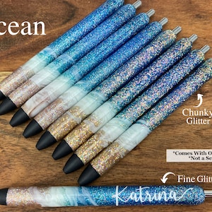 Puede incluir: Un conjunto de bol&iacute;grafos de purpurina azul y blanca con la palabra "Ocean" en la parte superior de la imagen. Los bol&iacute;grafos tienen un acabado de purpurina gruesa y fina. El texto "*Comes With One Pen *Not a Set" est&aacute; en la parte inferior de la imagen. La palabra "Katrina" est&aacute; escrita en cursiva en uno de los bol&iacute;grafos.