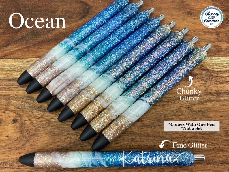 Puede incluir: Un conjunto de bol&iacute;grafos de purpurina azul y blanca con la palabra "Ocean" en la parte superior de la imagen. Los bol&iacute;grafos tienen un acabado de purpurina gruesa y fina. El texto "*Comes With One Pen *Not a Set" est&aacute; en la parte inferior de la imagen. La palabra "Katrina" est&aacute; escrita en cursiva en uno de los bol&iacute;grafos.