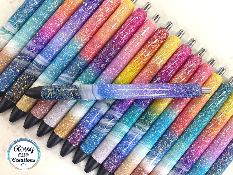 Puede incluir: Una colecci&oacute;n de 20 bol&iacute;grafos con purpurina con un degradado de colores, incluyendo morado, rosa, azul y amarillo. Los bol&iacute;grafos tienen una punta de tinta negra y una tapa de metal plateada. Los bol&iacute;grafos est&aacute;n dispuestos en forma de abanico sobre una superficie blanca. El texto "Glossy Cup Creations Co." es visible en la esquina inferior izquierda de la imagen.