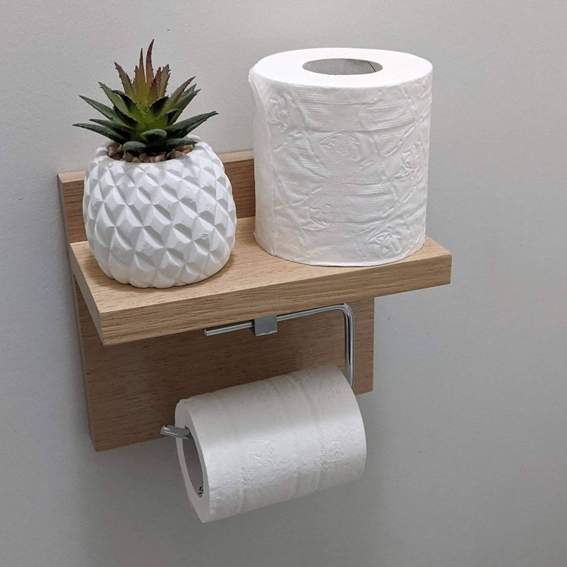 K&ouml;nnte beinhalten: Ein wandmontiertes Holzregal mit einem Toilettenpapierhalter und einem kleinen wei&szlig;en Topf mit einer gr&uuml;nen Sukkulente. Das Regal ist aus hellbraunem Holz und hat einen wei&szlig;en Keramiktopf mit Ananas-Design.