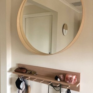 May include: A large, round wooden-framed mirror hangs above a wooden shelf with hooks. The shelf holds a brown case, sunglasses, and decorative items. Various items hang from the hooks, including a hat and bags. The wall is painted a light beige color.