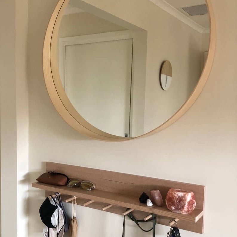 May include: A large, round wooden-framed mirror hangs above a wooden shelf with hooks. The shelf holds a brown case, sunglasses, and decorative items. Various items hang from the hooks, including a hat and bags. The wall is painted a light beige color.