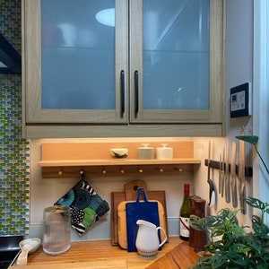May include: A kitchen scene featuring wooden cabinets with frosted glass doors, a wooden shelf with small bowls and containers, and a wooden countertop with cutting boards, a pitcher, and a wine bottle. A green plant is in the foreground.