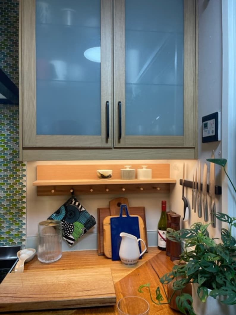 May include: A kitchen scene featuring wooden cabinets with frosted glass doors, a wooden shelf with small bowls and containers, and a wooden countertop with cutting boards, a pitcher, and a wine bottle. A green plant is in the foreground.