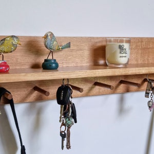 May include: A wooden key holder with a shelf, featuring decorative bird figurines, a candle, and a metal heart. Below the shelf are several pegs holding keys and a black bag. The key holder is mounted on a white wall.