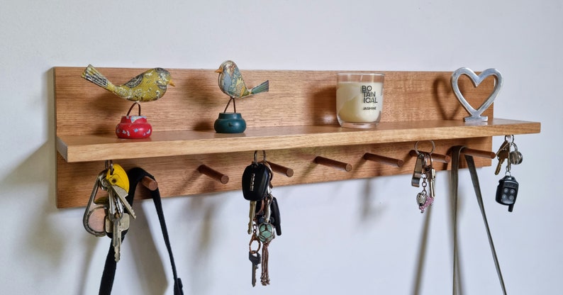 May include: A wooden key holder with a shelf, featuring decorative bird figurines, a candle, and a metal heart. Below the shelf are several pegs holding keys and a black bag. The key holder is mounted on a white wall.