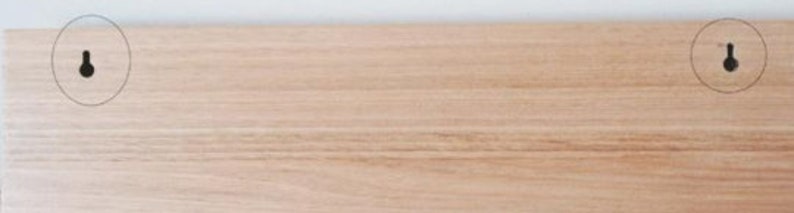 May include: A light-colored wooden board with two black keyhole hangers. The wood grain is visible, and the board is mounted on a white wall. The hangers are evenly spaced.