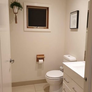 K&ouml;nnte beinhalten: Eine wei&szlig;e Toilette mit einem wei&szlig;en Toilettensitz in einem Badezimmer mit wei&szlig;en W&auml;nden und einem wei&szlig;en Waschtisch mit Schubladen. &Uuml;ber der Toilette befindet sich ein kleines Fenster und an der Wand h&auml;ngt ein gerahmtes Bild.