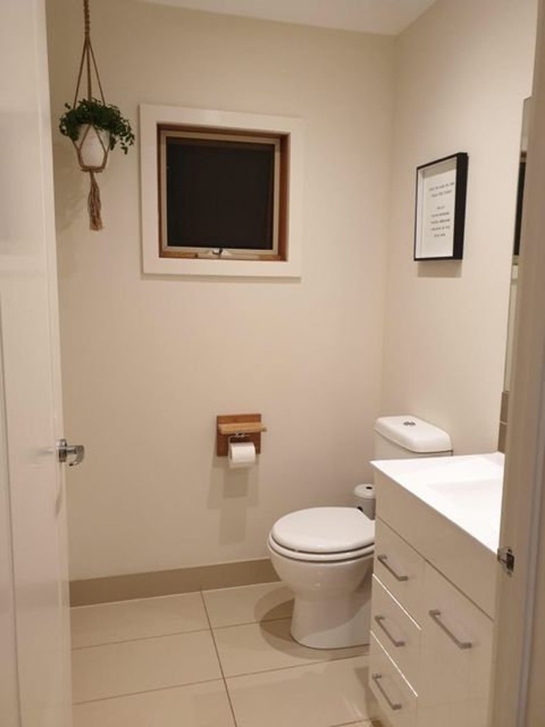 K&ouml;nnte beinhalten: Eine wei&szlig;e Toilette mit einem wei&szlig;en Toilettensitz in einem Badezimmer mit wei&szlig;en W&auml;nden und einem wei&szlig;en Waschtisch mit Schubladen. &Uuml;ber der Toilette befindet sich ein kleines Fenster und an der Wand h&auml;ngt ein gerahmtes Bild.