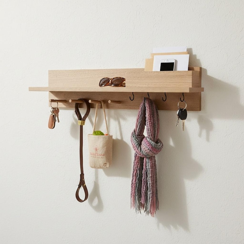 May include: A light-colored wooden wall-mounted organizer with a shelf, hooks, and a small compartment. It holds sunglasses, keys, a dog leash, a small bag, a scarf, and a phone. The organizer is mounted on a white wall.