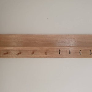 May include: A wooden key holder with a shelf and pegs. The key holder is made of light-colored wood and has a rectangular shelf at the top. Below the shelf are four wooden pegs and four black metal hooks.