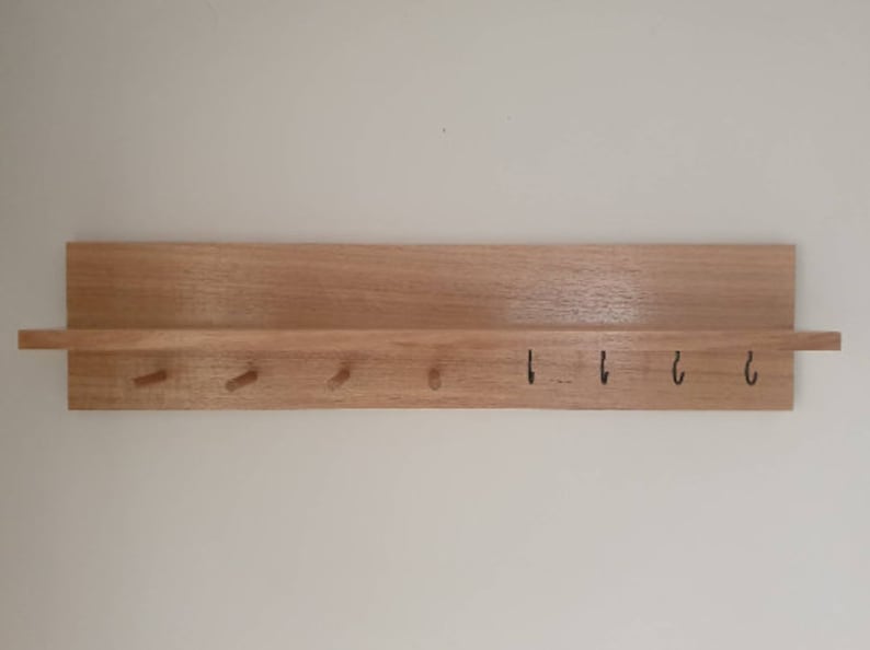 May include: A wooden key holder with a shelf and pegs. The key holder is made of light-colored wood and has a rectangular shelf at the top. Below the shelf are four wooden pegs and four black metal hooks.