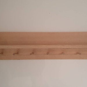 May include: A wooden wall shelf with a small shelf and six pegs. The shelf is made of light-colored wood and is mounted on a white wall. The pegs are evenly spaced below the shelf.