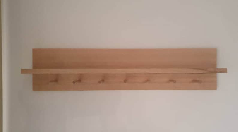 May include: A wooden wall shelf with a small shelf and six pegs. The shelf is made of light-colored wood and is mounted on a white wall. The pegs are evenly spaced below the shelf.