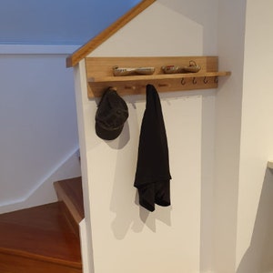 May include: A wooden wall-mounted coat rack with a shelf, hooks, and pegs. A black cap and coat hang from the rack. Two small metal trays sit on the shelf. The rack is mounted on a white wall near a wooden staircase.