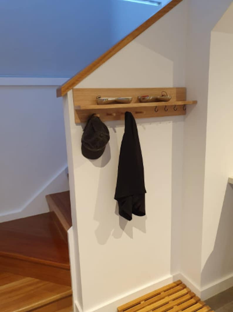 May include: A wooden wall-mounted coat rack with a shelf, hooks, and pegs. A black cap and coat hang from the rack. Two small metal trays sit on the shelf. The rack is mounted on a white wall near a wooden staircase.
