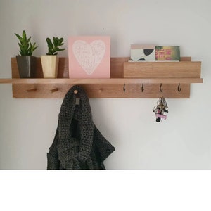May include: A wooden wall shelf with a light brown finish, featuring a top shelf with small potted plants and a pink square with white text. Below are hooks for hanging items, with a scarf and keys visible.